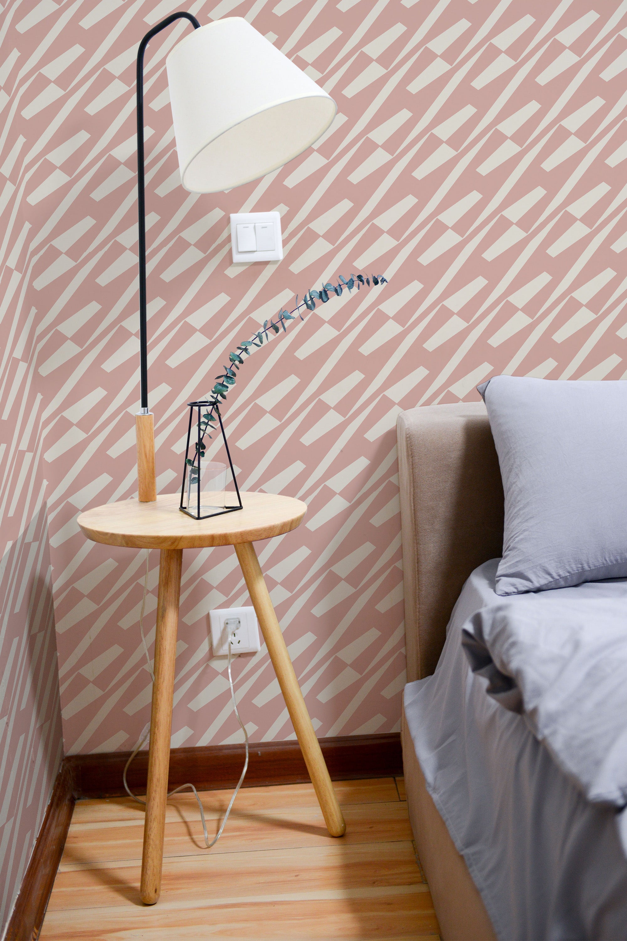 Self-adhesive wallpaper with a geometric herringbone pattern in soft pink and white tones on a room wall beside a modern floor lamp and wooden side table.