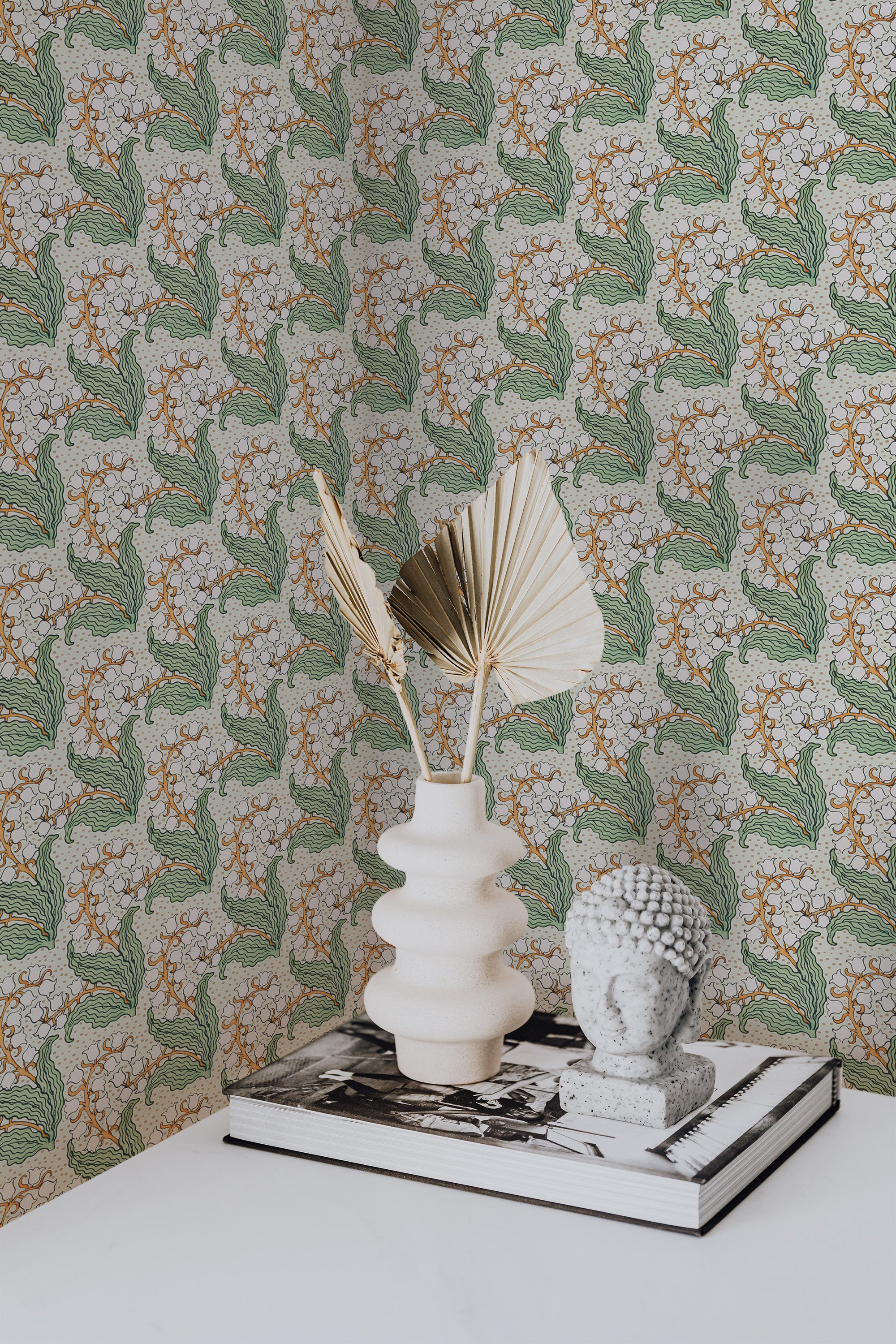 Self-adhesive wallpaper with a repeating pattern of Lilies of the Valley on a backdrop, accompanied by decorative items and a book on a surface.