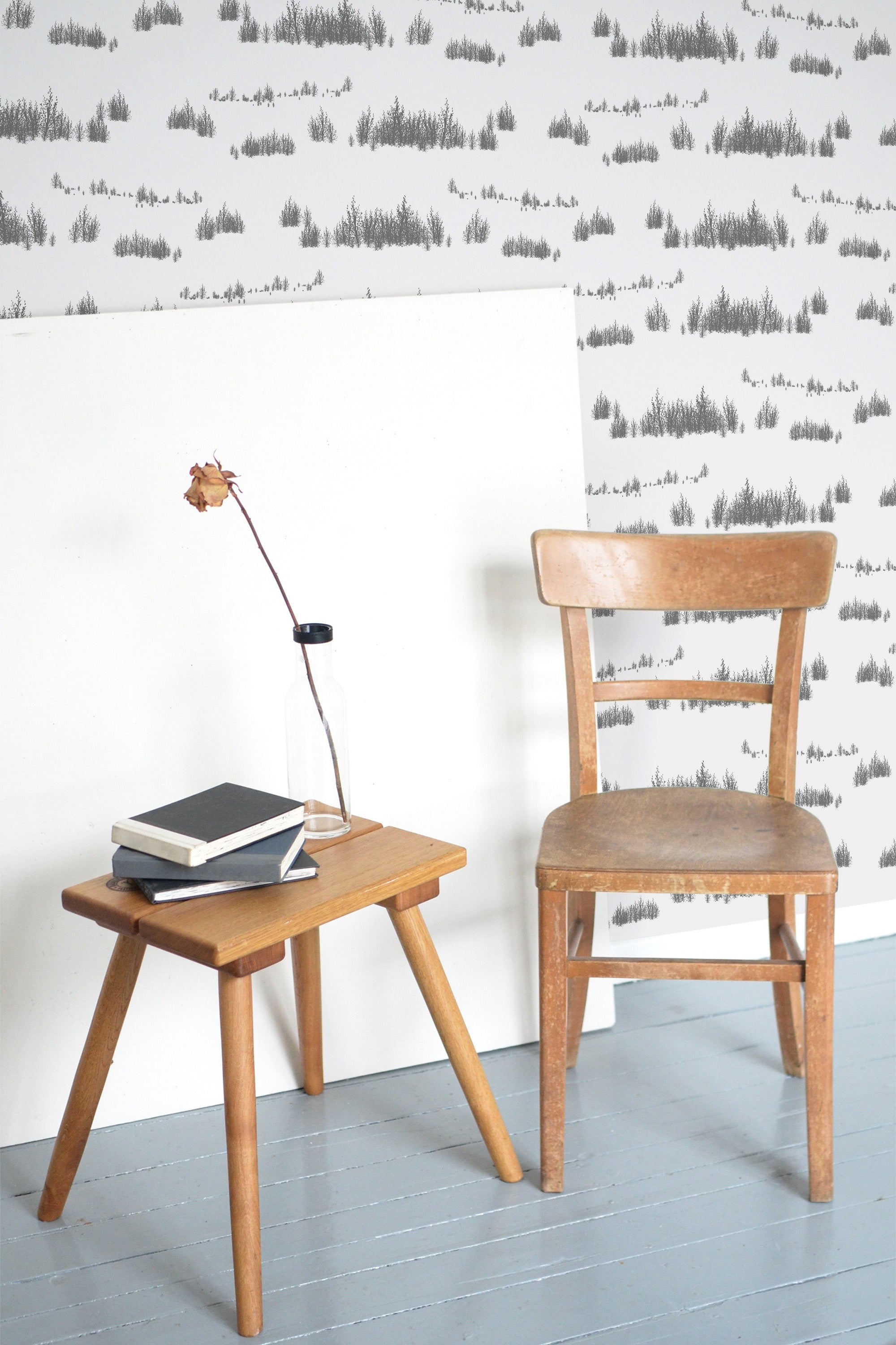 Self-adhesive wallpaper with a repeating pattern of gray trees on a light background, accented by a wooden chair and side table with books and a lamp.