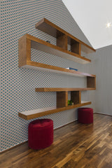 A room with geometric pink and green Art Deco wallpaper, accompanied by wooden shelving and two red ottomans, on a herringbone parquet floor, traditional wallpaper.