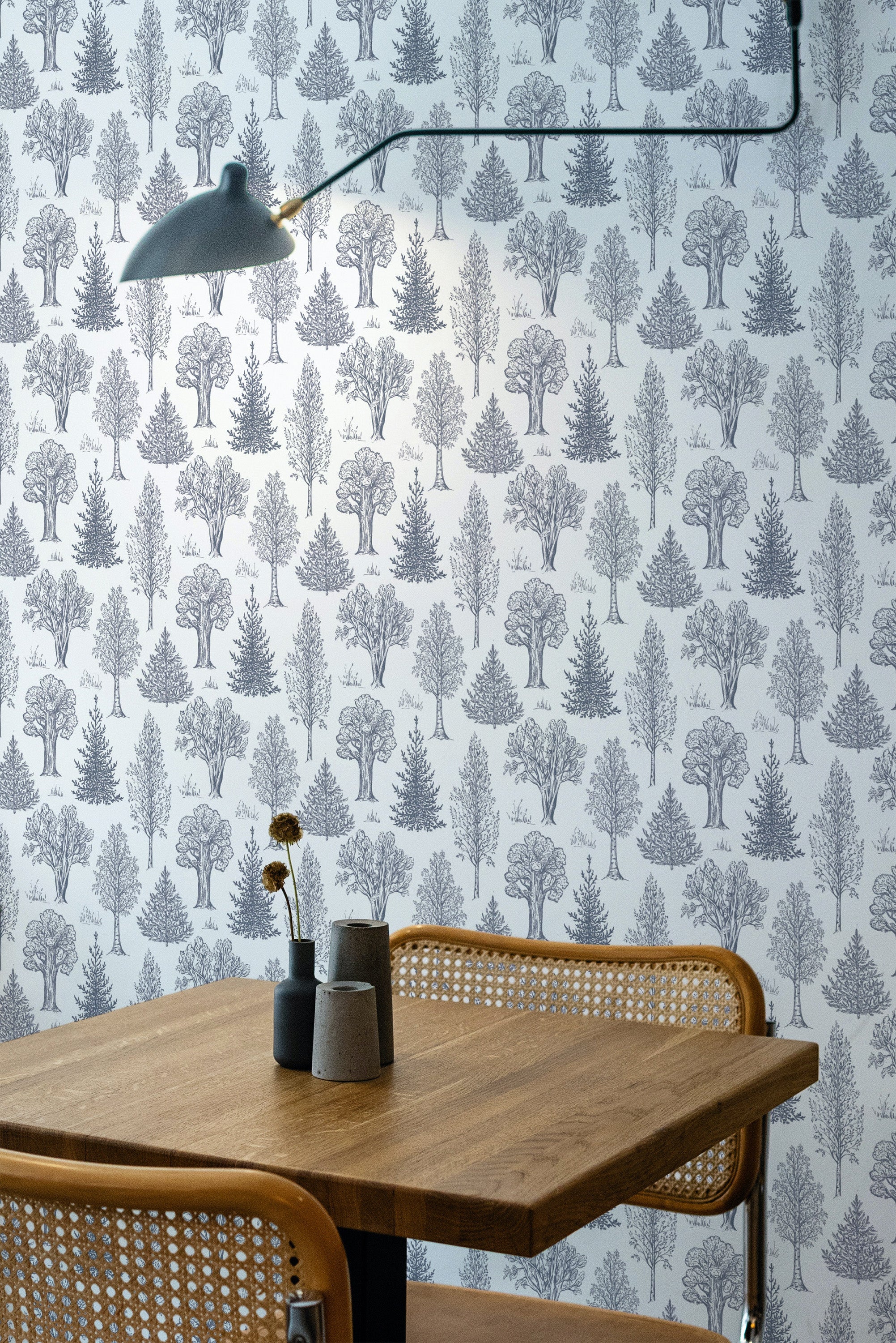 Self-adhesive wallpaper with a repeating pattern of delicate trees in a light aesthetic design, covering a room's wall behind a wooden table.