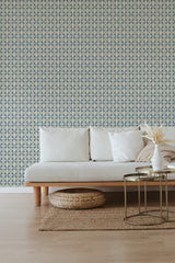 A room with one wall covered in Busy Tile wallpaper with a repetitive blue and beige geometric pattern, alongside a neutral-toned couch and a woven ottoman, embodying a traditional wallpaper.