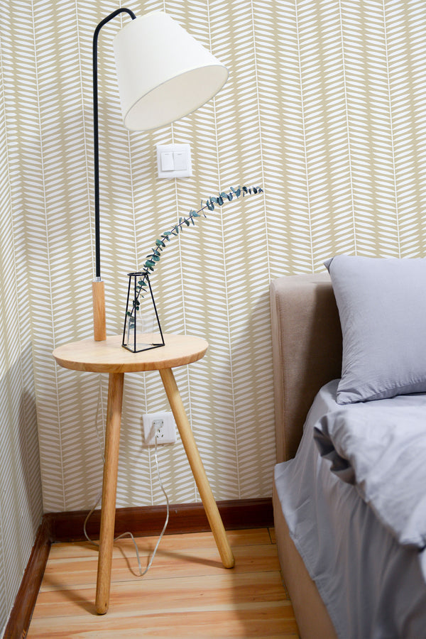 Self-adhesive Honey Herringbone pattern wallpaper on a bedroom wall with a light wooden side table, a lamp, and a bed with grey bedding.