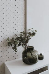 Self-adhesive wallpaper with a Tiny Cross pattern in black on a white background, affixed to a room wall, complemented by a green vase with leaves on a white shelf.