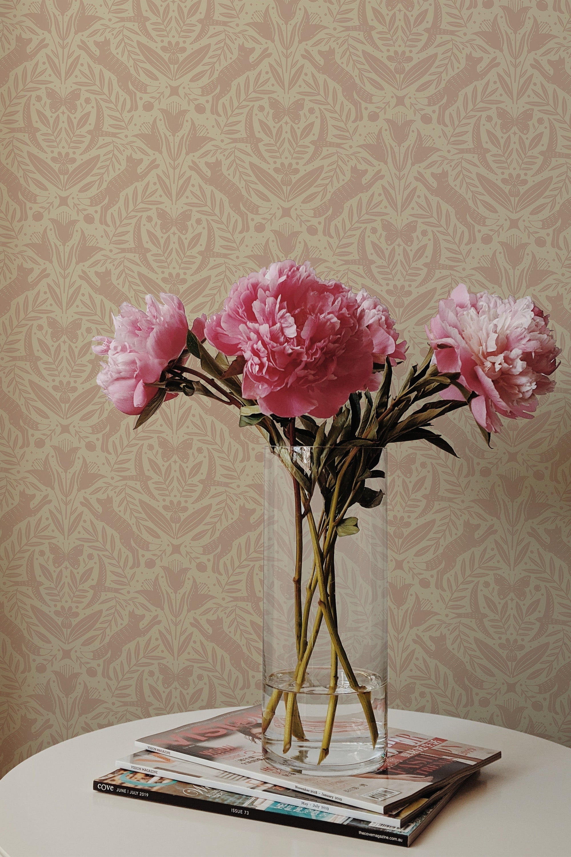 Self-adhesive wallpaper with an elegant woodland-inspired pattern on a room wall, accented by a vase of pink peonies on a stack of magazines.