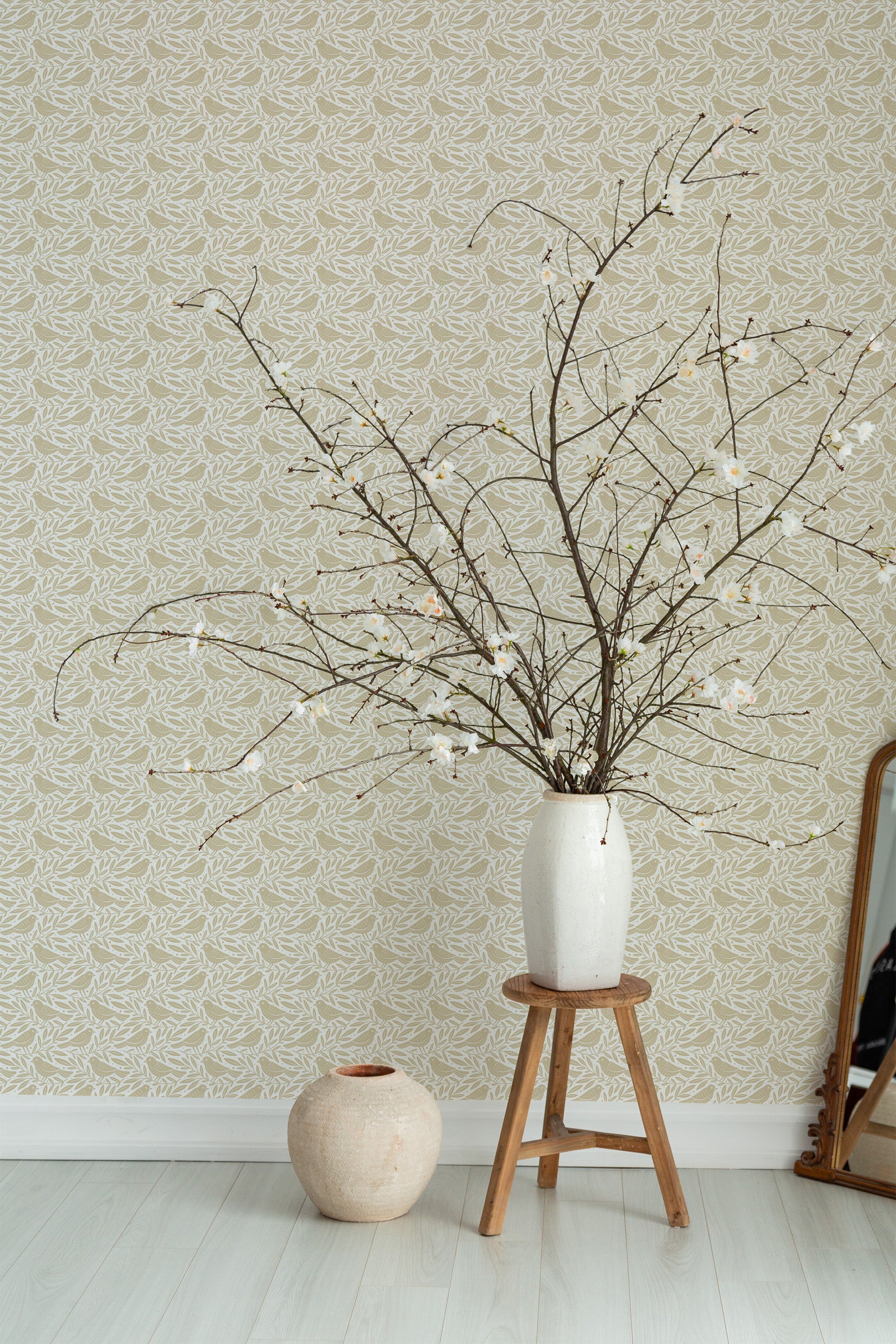 Self-adhesive Honey Bird wallpaper with a repeating pattern of stylized birds and floral motifs in soft beige tones on a muted background, complemented by a vase with branches on a wooden stool.