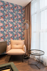 A room with a wall covered in Tropical Bird wallpaper with a pattern of colorful birds and foliage on a blue background, alongside a cream chair and table, near sheer-curtained windows, traditional wallpaper.