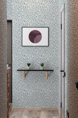 A room with Rainstorm brush lines design wallpaper with swirling gray patterns on a lighter background, complemented by a framed geometric artwork, a shelf with plants, and a traditional wallpaper finish.