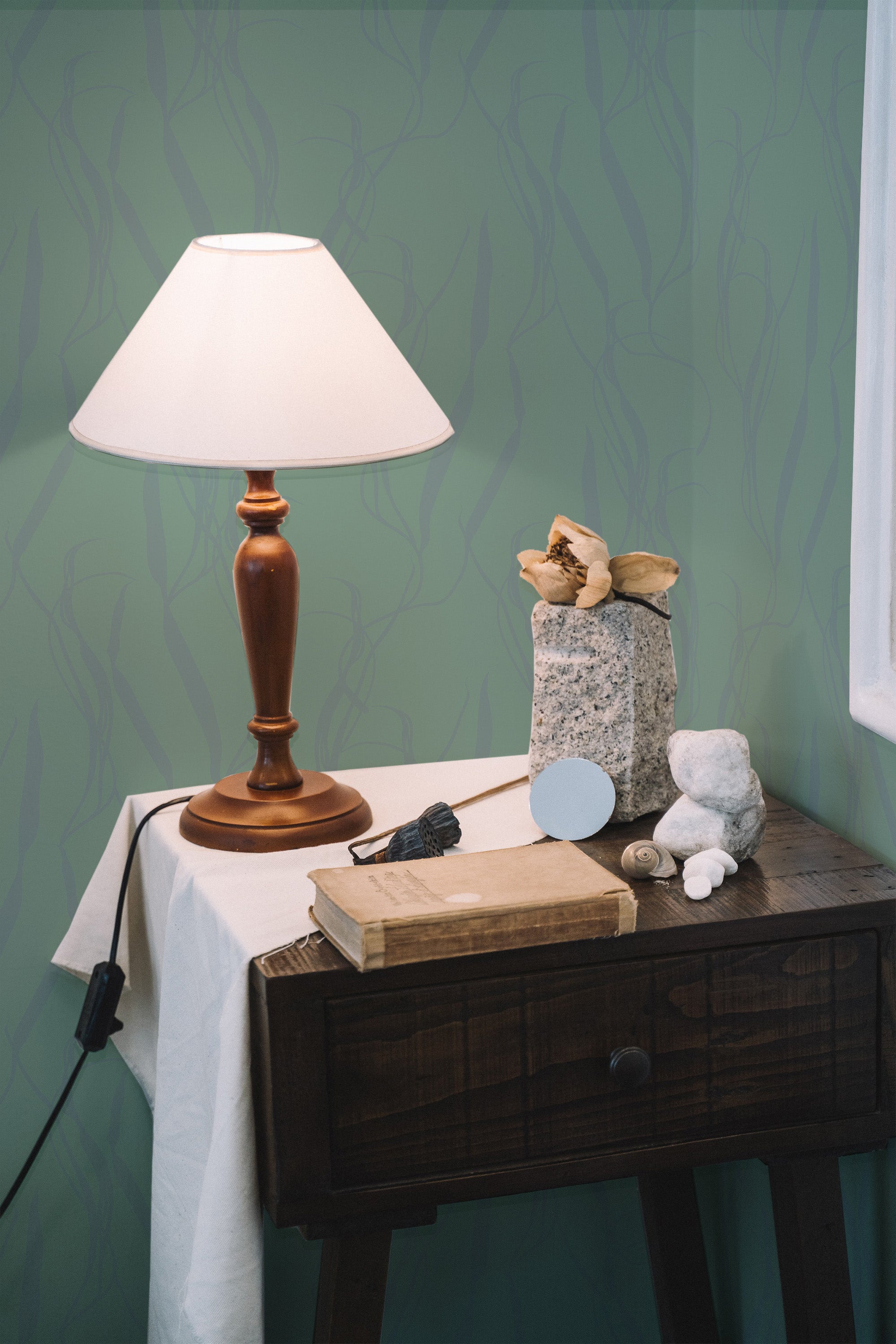 Self-adhesive wallpaper with an aesthetic leaf sage design on a room wall, complemented by a wooden side table, a lamp, and decorative items.