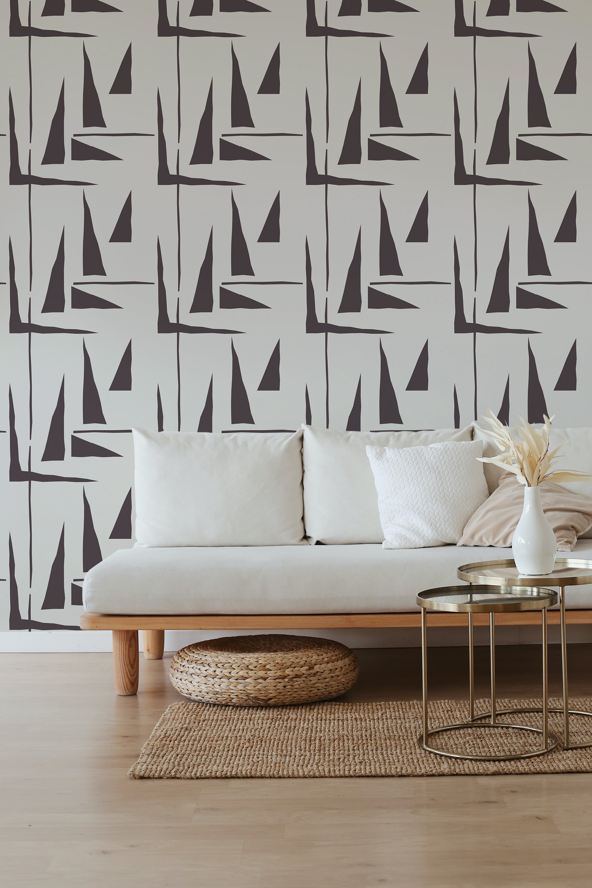 A room with a wall covered in 'Simple triangles' patterned wallpaper, which includes varying sizes of black triangles on a cream background, creating a modern geometric look, behind a beige sofa, traditional wallpaper.