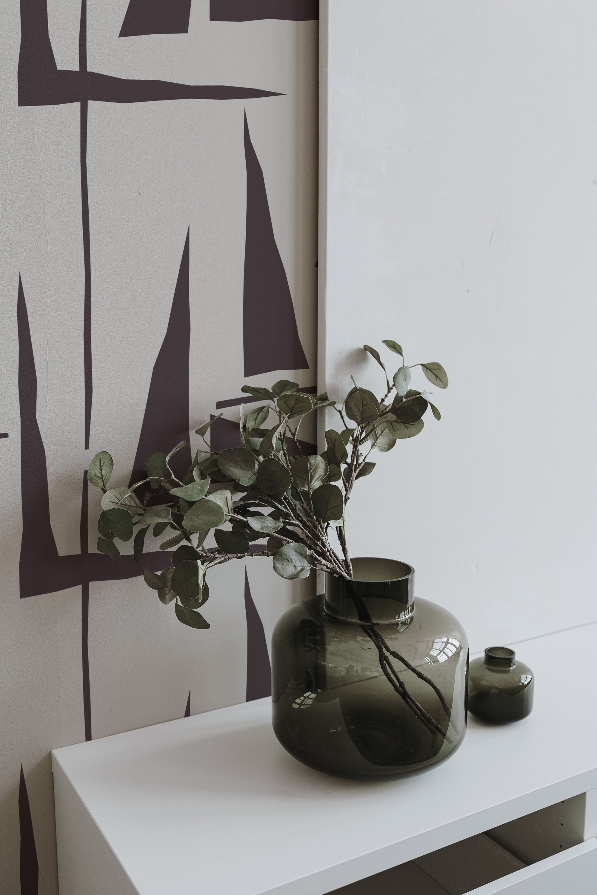 Self-adhesive wallpaper with a geometric pattern of simple triangles in various shades of purple on a wall, with a shelf holding a green vase and branches.