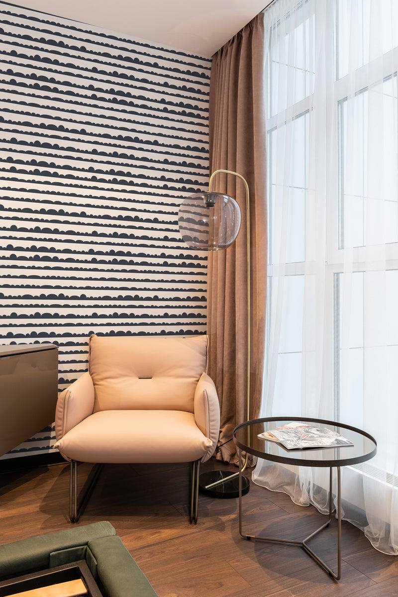 A cozy room corner with a wall covered in Graphic Cloud Line wallpaper, consisting of undulating black lines on a white background, complemented by a tan armchair and curtains, traditional wallpaper.