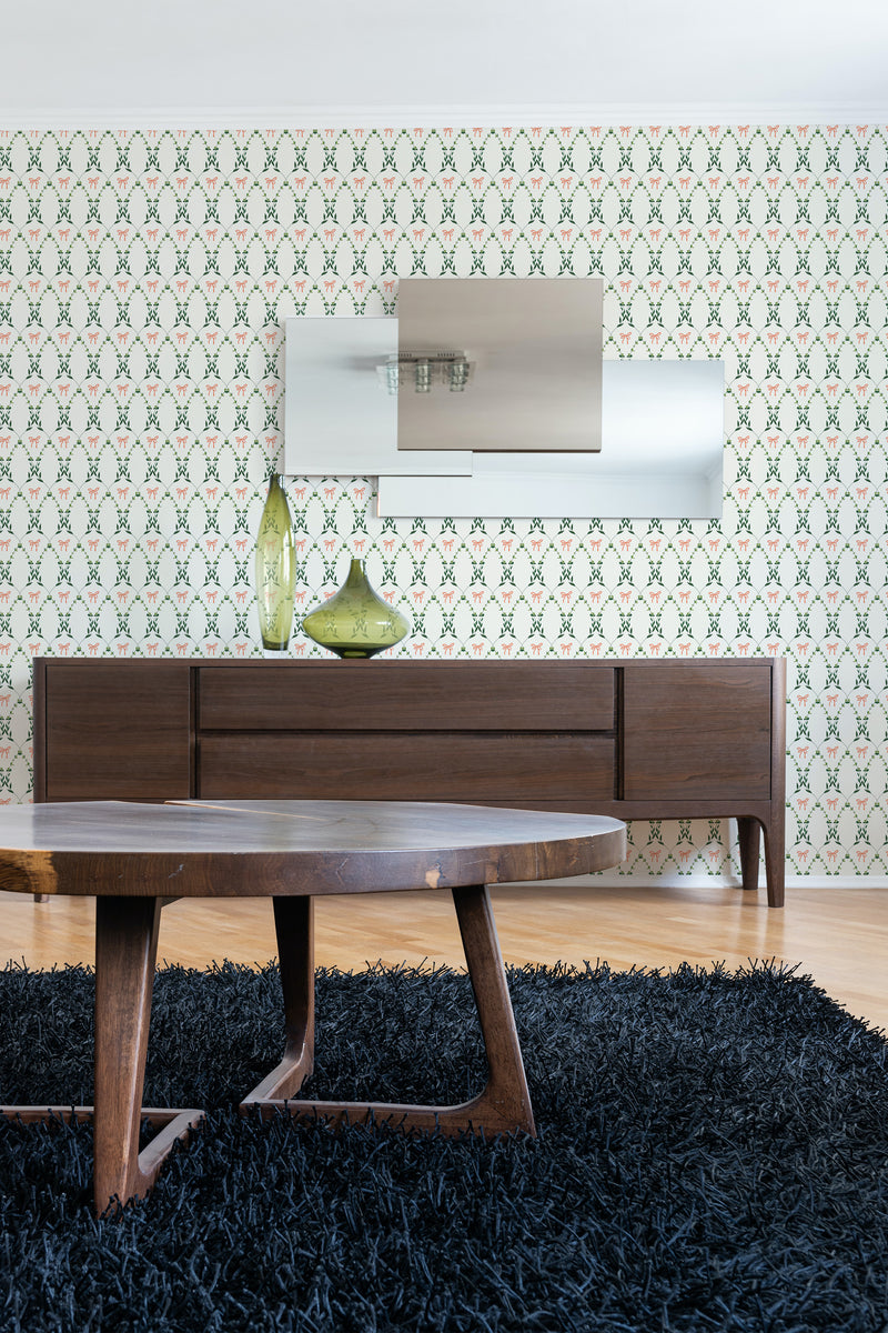 A room with a wall covered in Country Floral Bow wallpaper with a pattern of green bows and small flowers on a cream background, above a wooden sideboard and a dark blue rug, traditional wallpaper.