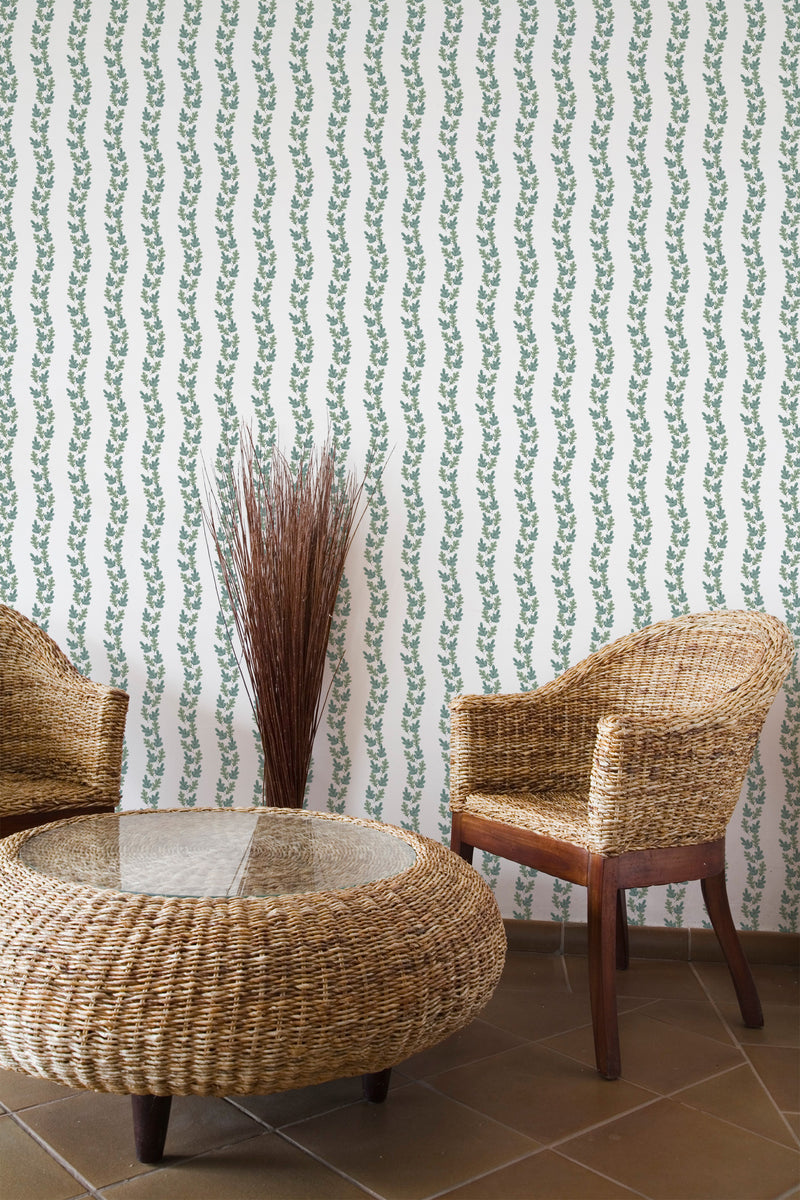 Self-adhesive wallpaper with green and white wavy vines pattern on a room wall, with wicker furniture and decorative twigs.