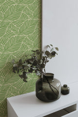 Self-adhesive Green Vintage Leaves wallpaper with a pattern of overlapping foliage in shades of green, applied to a wall beside a white canvas and green vases with branches.