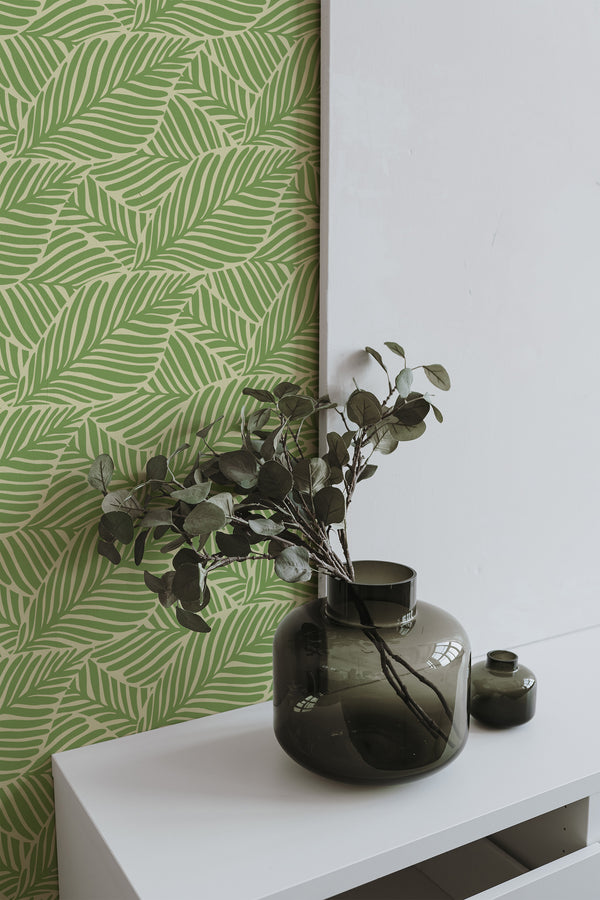 Self-adhesive Green Vintage Leaves wallpaper with a pattern of overlapping foliage in shades of green, applied to a wall beside a white canvas and green vases with branches.