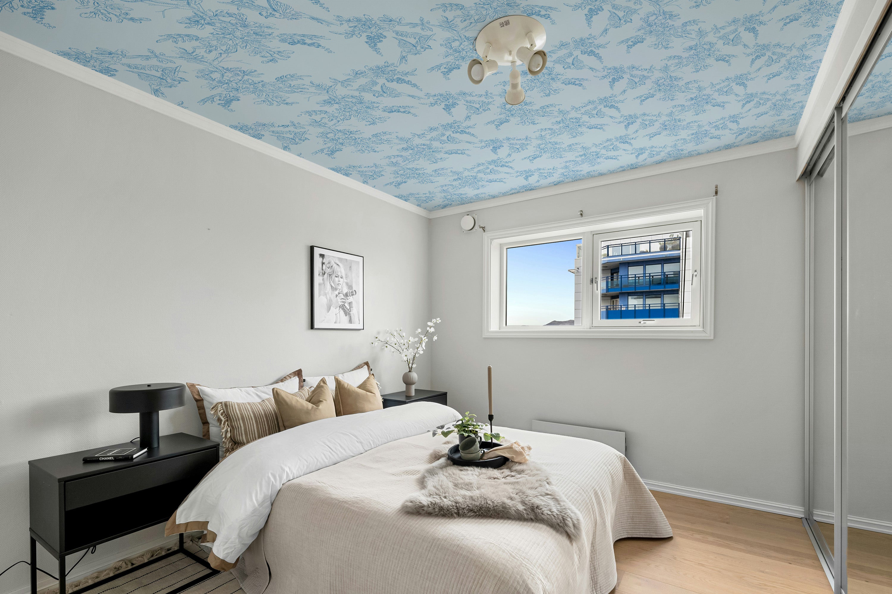 Modern bedroom with a decorative blue ceiling pattern, bed with pillows, nightstands, and a window.