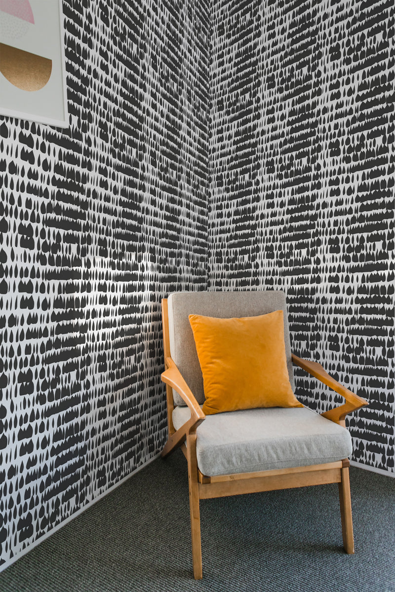 The wallpaper has a black-and-white paint blot pattern with irregular, vertical blotches spread uniformly across the wall. The design creates a bold, textured aesthetic. On one side of the room, a modern wooden chair with a mustard-colored cushion sits against the wall. Traditional wallpaper.