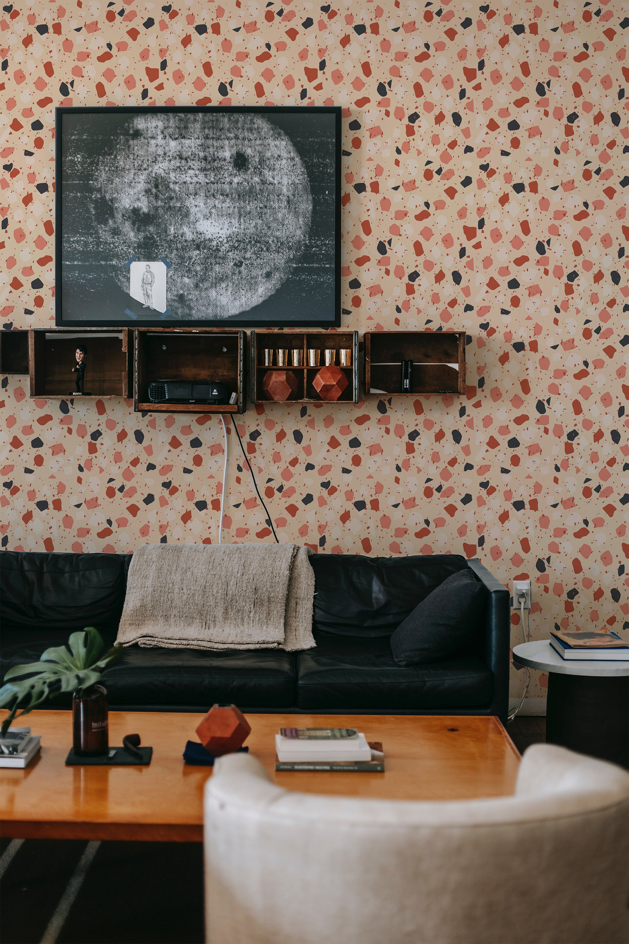 A room with a Terrazzo design wallpaper featuring scattered, irregular shapes in earthy tones of terracotta, beige, black, and gray against a light background. The wallpaper is installed behind a black leather couch, wooden floating shelves, and artwork, contributing to a warm and textured look. Traditional wallpaper.