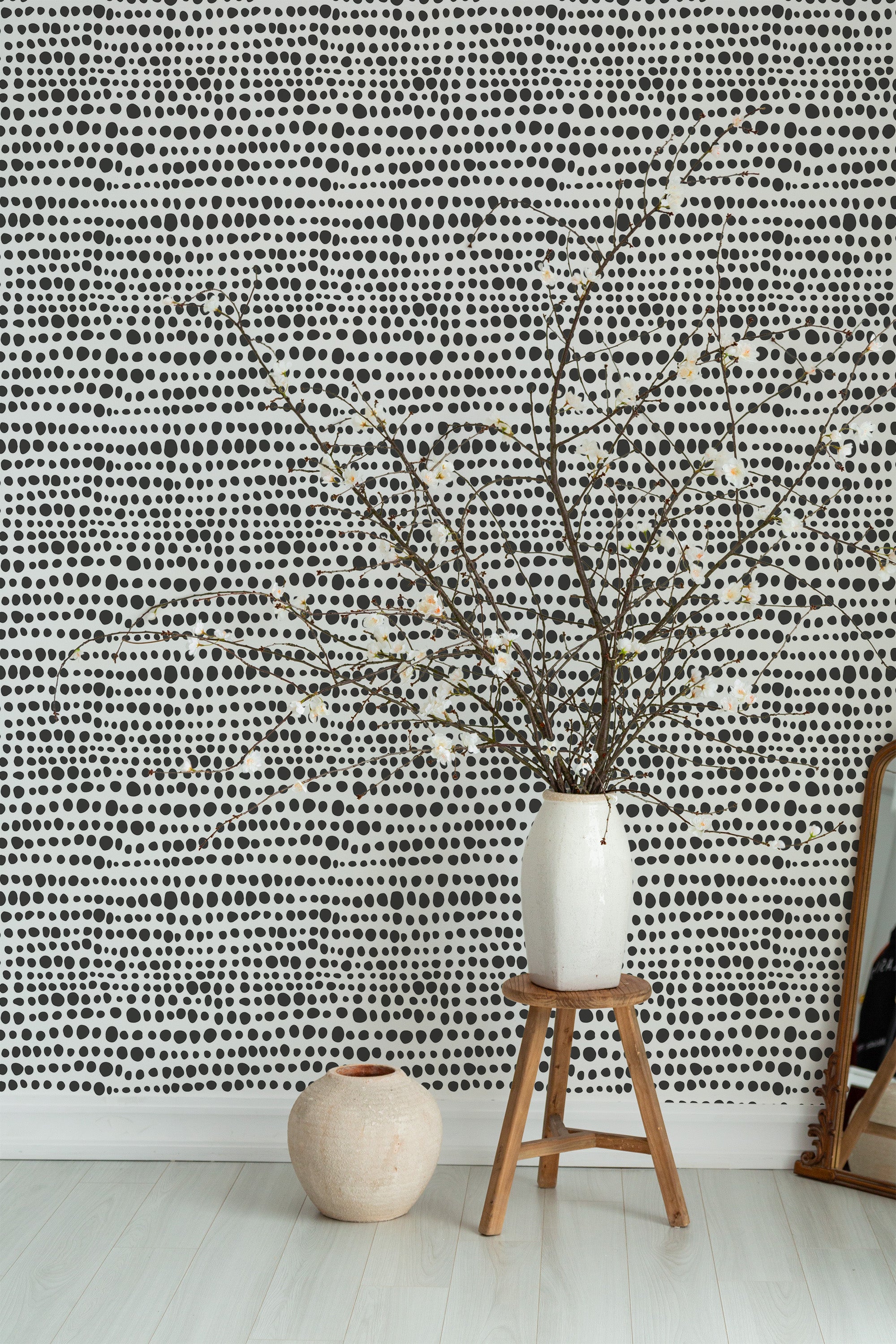 Self-adhesive wallpaper with a black and white polka dot pattern, arranged in a wavy, irregular grid. The design adds a modern and playful touch to the room, complementing the minimalist decor with a stool, ceramic vases, and a leaning mirror.