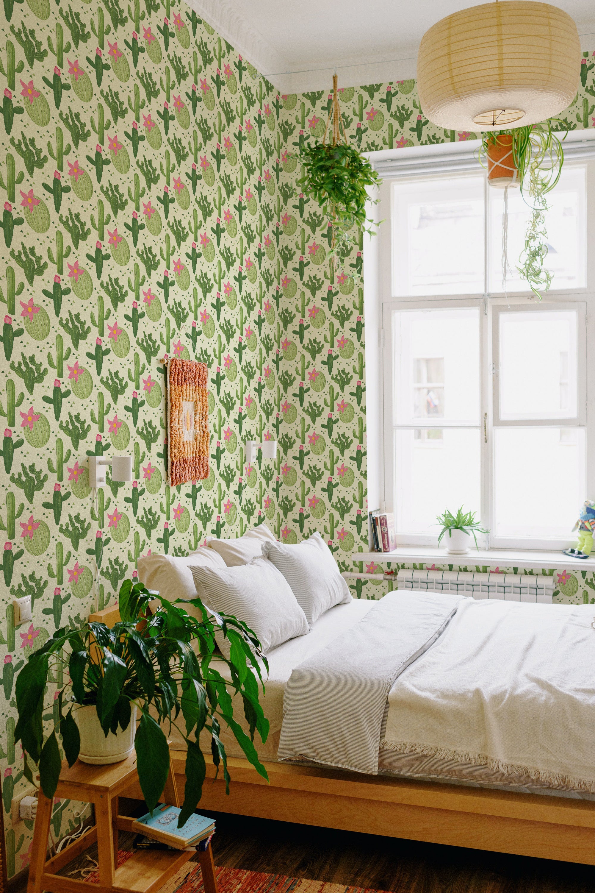Wallpaper with a repeating cactus pattern in shades of green and pink accents, covering the entire wall around a bed with natural light coming from a large window. The design creates a lively, nature-inspired atmosphere in the room. Traditional wallpaper.