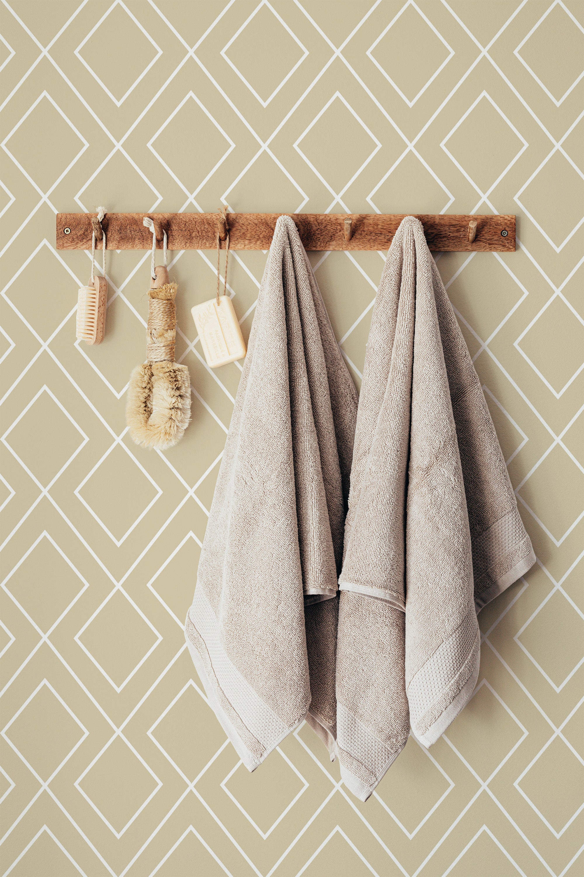 Self-adhesive geometric wallpaper with a minimalist design of white diamonds on a beige background covering the wall behind a wooden rack holding towels and bath accessories.
