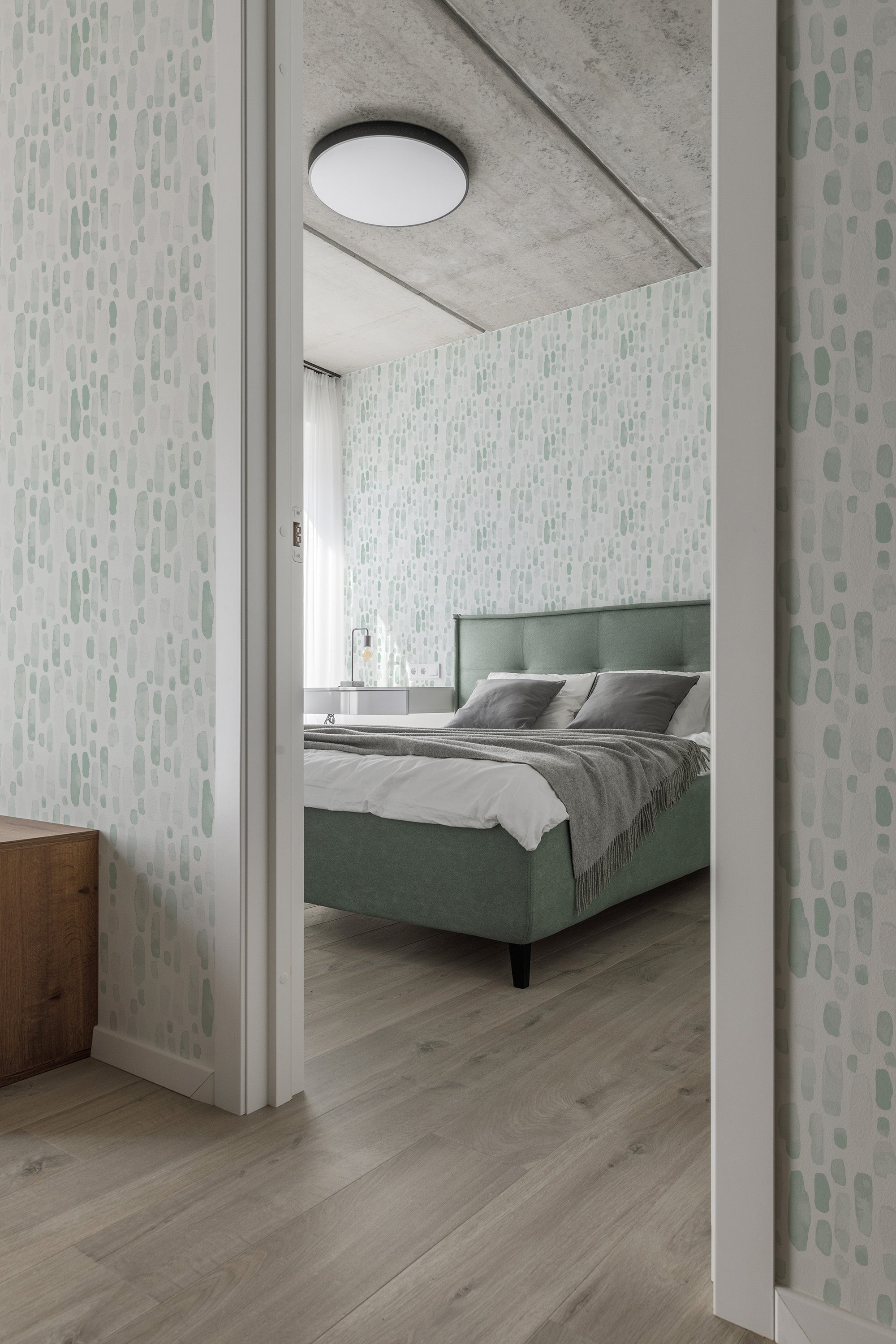A room with walls covered in green watercolor drops, creating a soft and calming atmosphere. The abstract pattern consists of various shades of green in irregular oval shapes spread evenly across a light background. Traditional wallpaper.