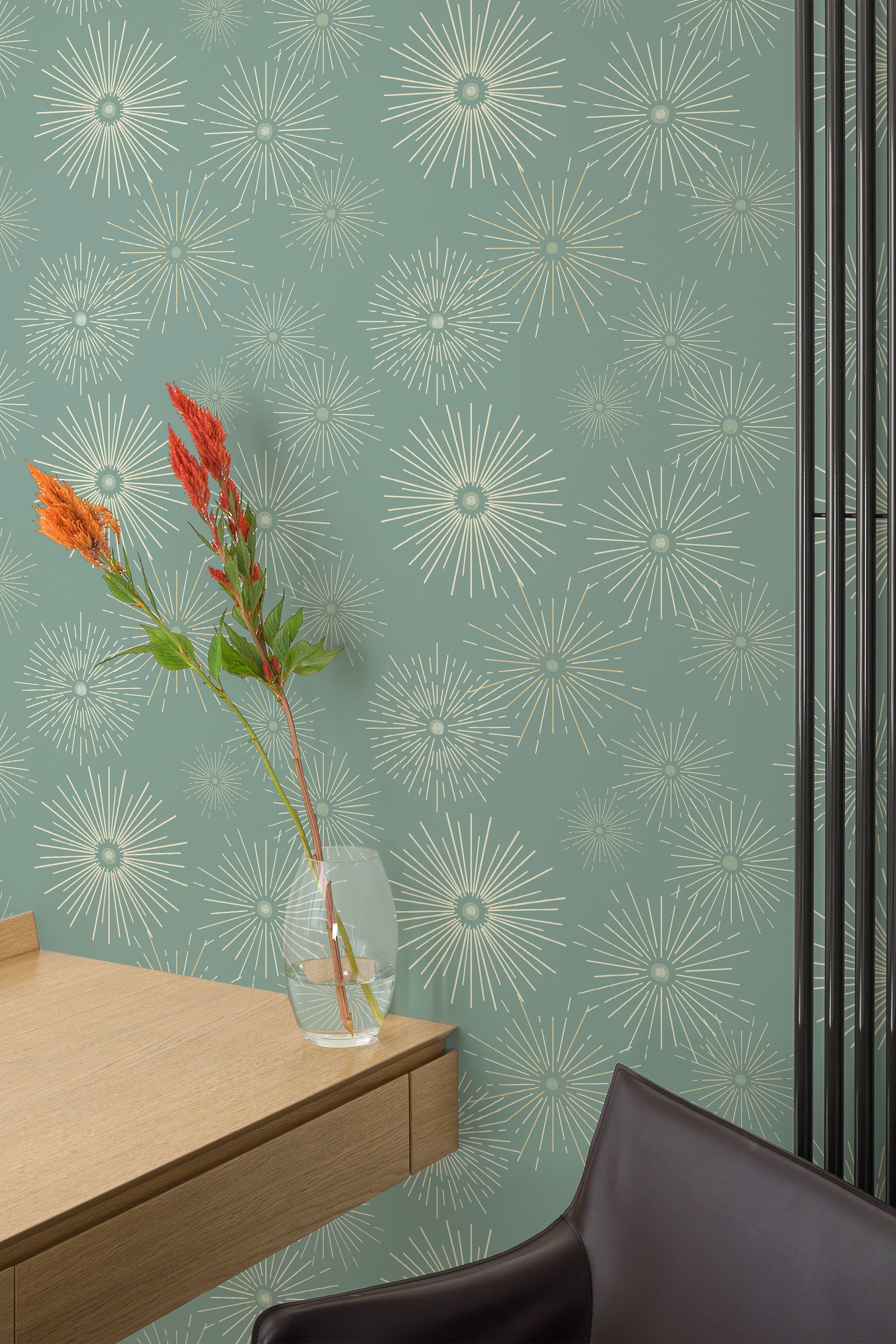 Self-adhesive wallpaper with a Green starburst design on a muted sage green background, paired with a single glass vase containing red and orange flowers next to a wooden desk and brown chair.