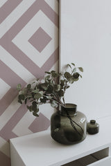 Self-adhesive wallpaper with a geometric square design in soft, muted tones covering one wall, accompanied by a minimalist shelf holding green glass vases with eucalyptus branches.