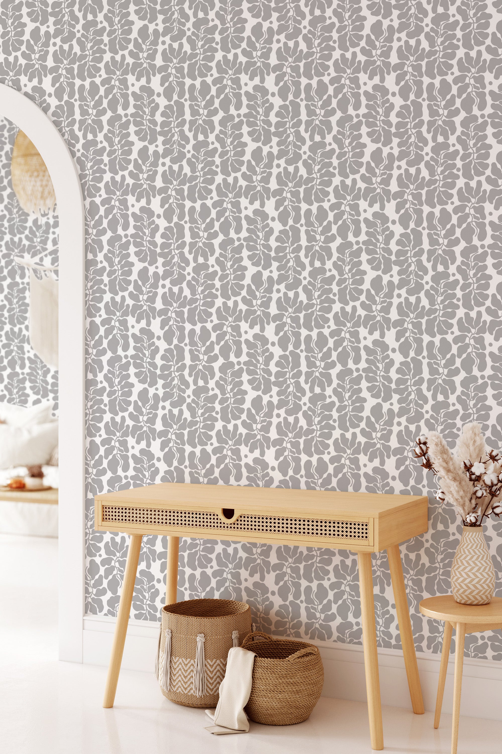 A room with a wall covered in abstract leaf-patterned wallpaper, blending various shades of gray on a white background, complemented by a wooden console table, woven baskets, and decorative vases, embodying a modern twist on traditional wallpaper.