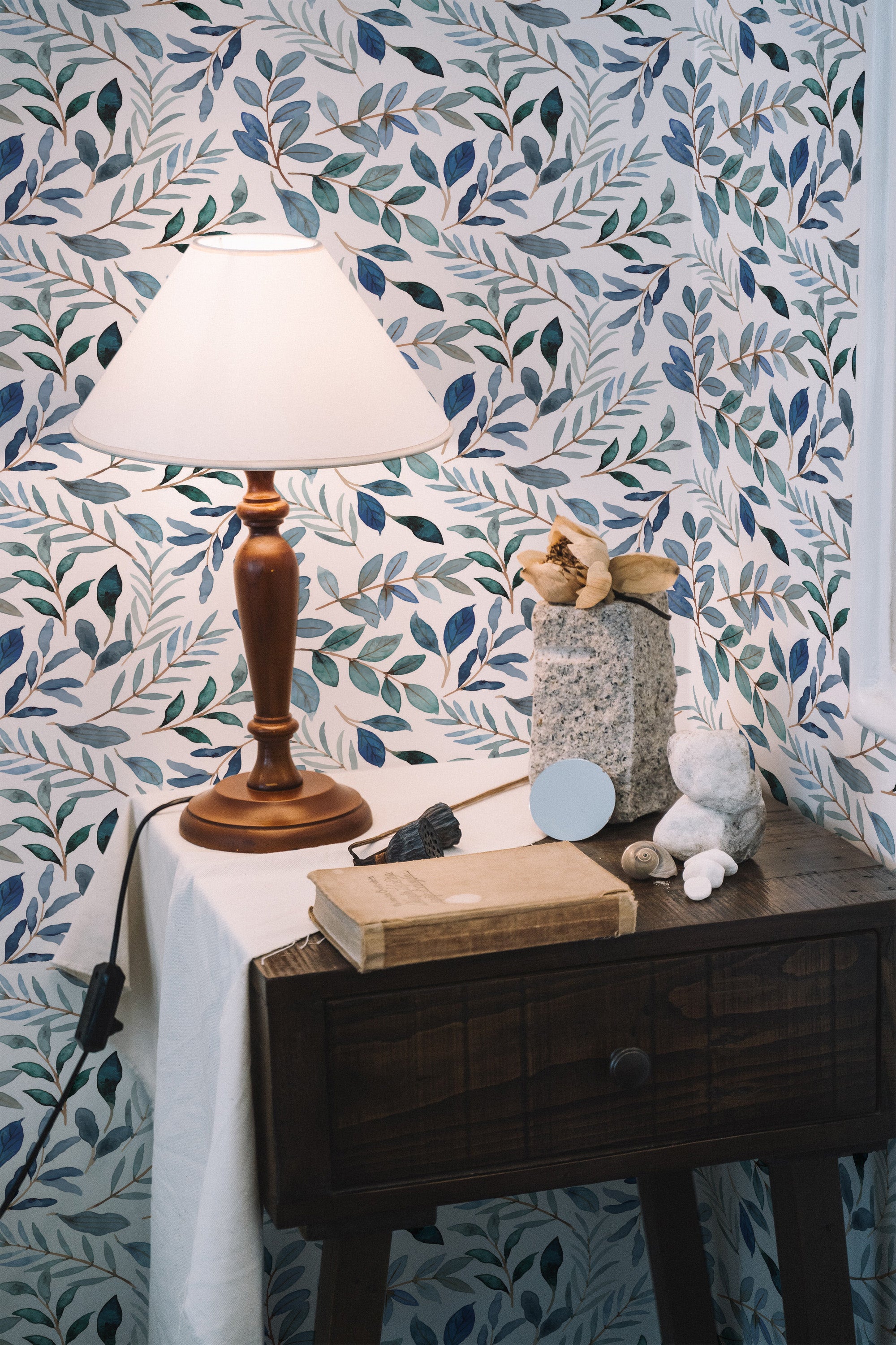 Self-adhesive blue watercolor leaf wallpaper in a room with a wooden table, lamp, and decorative items.