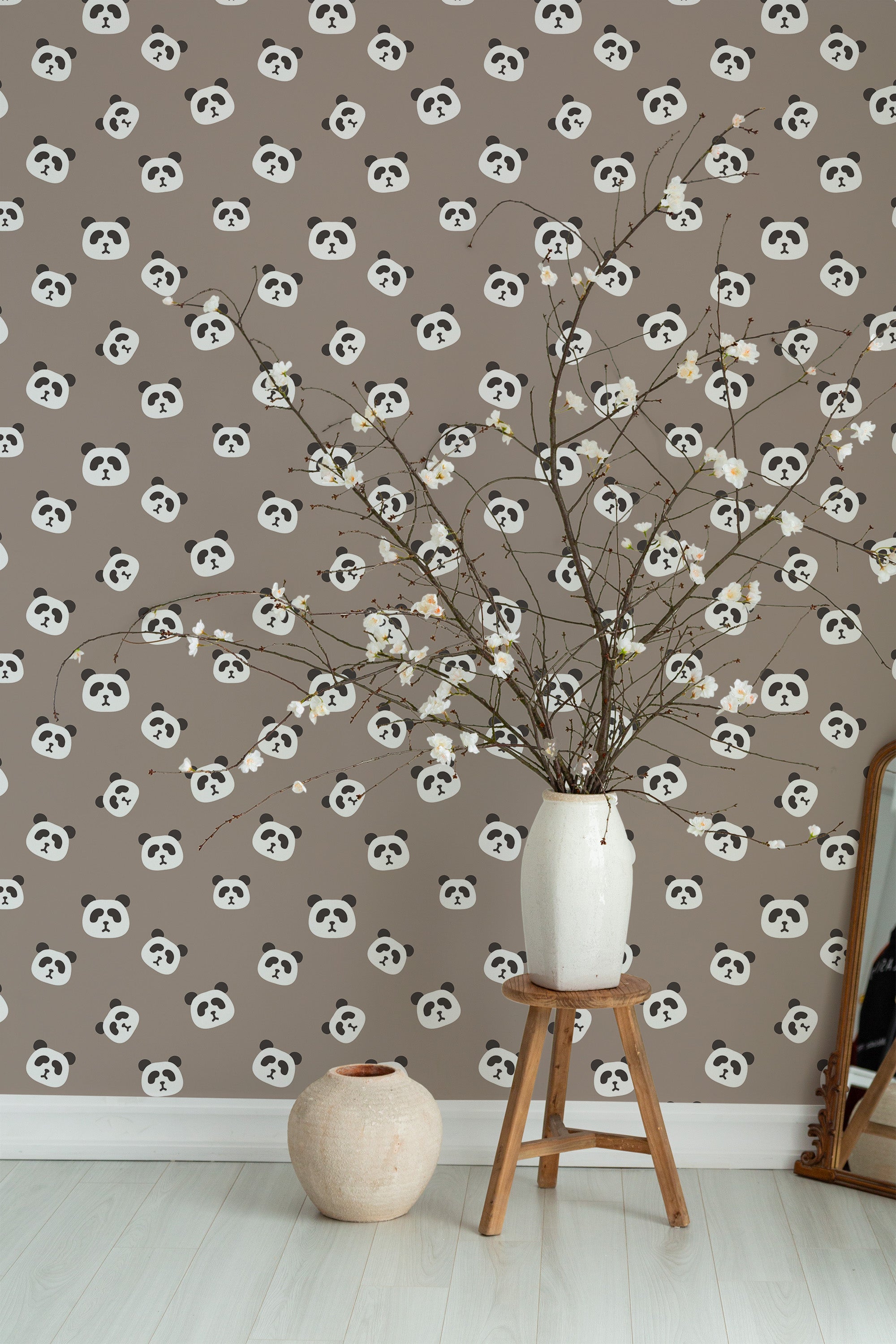 Self-adhesive wallpaper with a repeating pattern of panda faces on a grey background, accented by a vase of branches on a wooden stool.