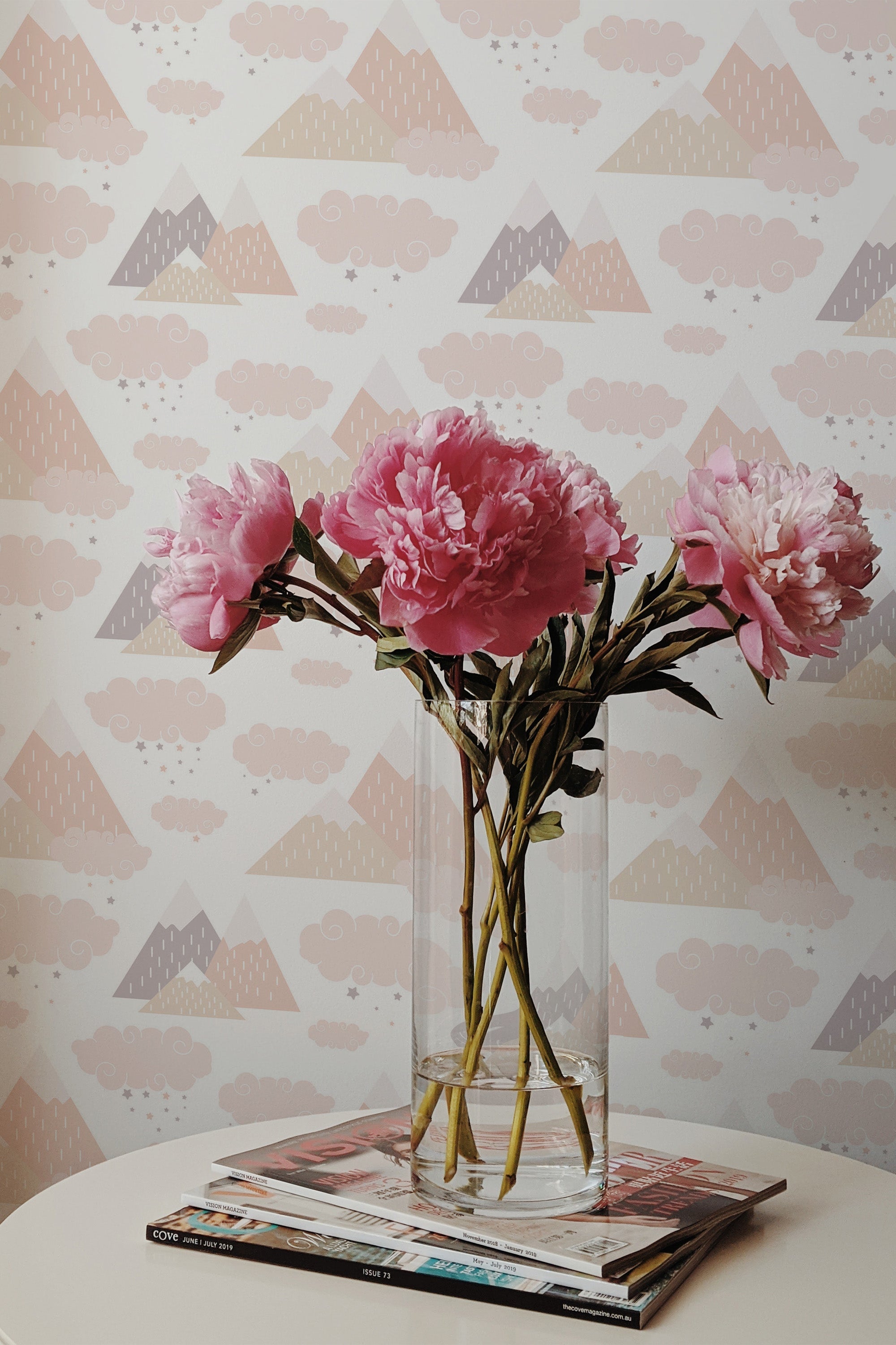 Self-adhesive wallpaper with geometric mountain patterns in shades of pink, gray, and beige, complemented by cloud motifs, installed in a stylish room.
