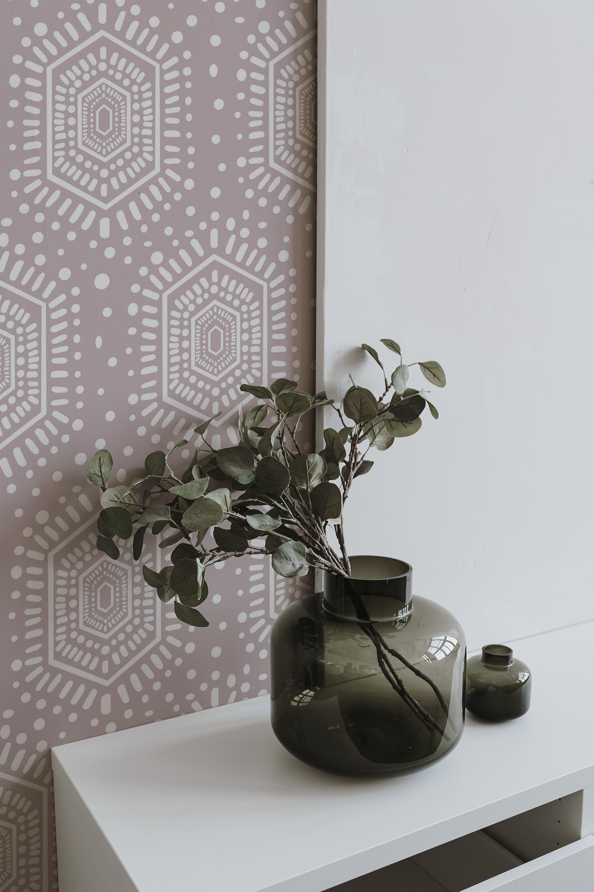 Self-adhesive ethnic patterned wallpaper with geometric shapes on a soft pink background, accompanied by decorative green vases and foliage on a white shelf.