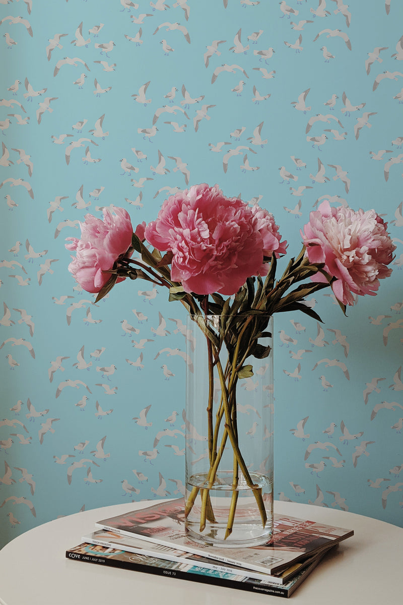 Self-adhesive wallpaper with a pattern of flying seagulls on a light blue background, complemented by a vase of pink peonies on a table.