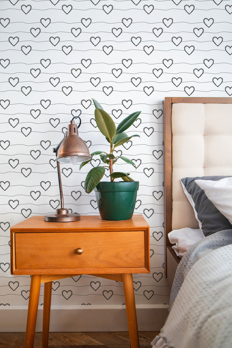 Self-adhesive wallpaper with repeated black heart line art pattern on a white background in a cozy bedroom setting with a wooden bedside table, table lamp, and potted plant.