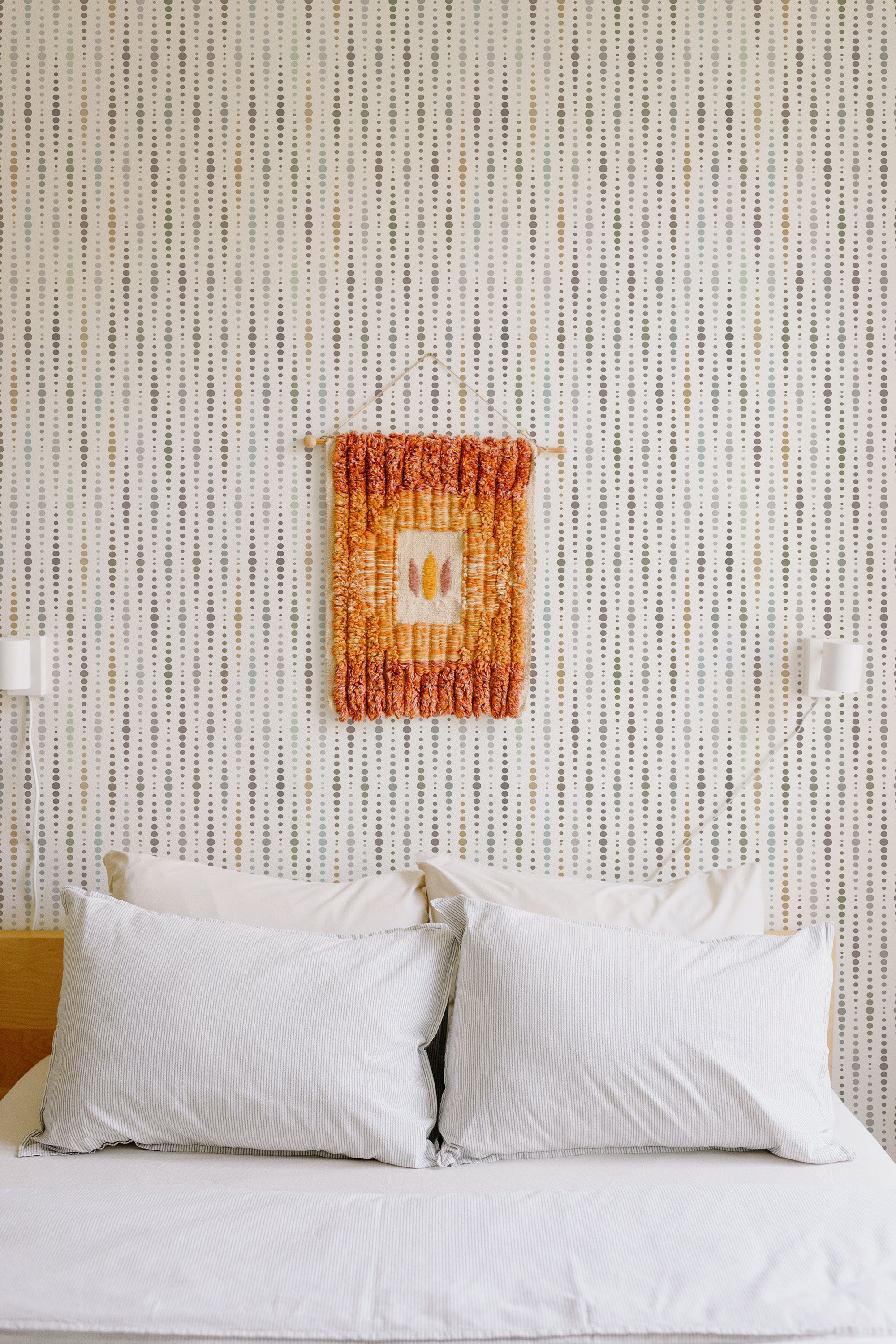 Self-adhesive wallpaper with a neutral dots pattern on a room's wall, above a bed with white pillows and beside white wall lamps.