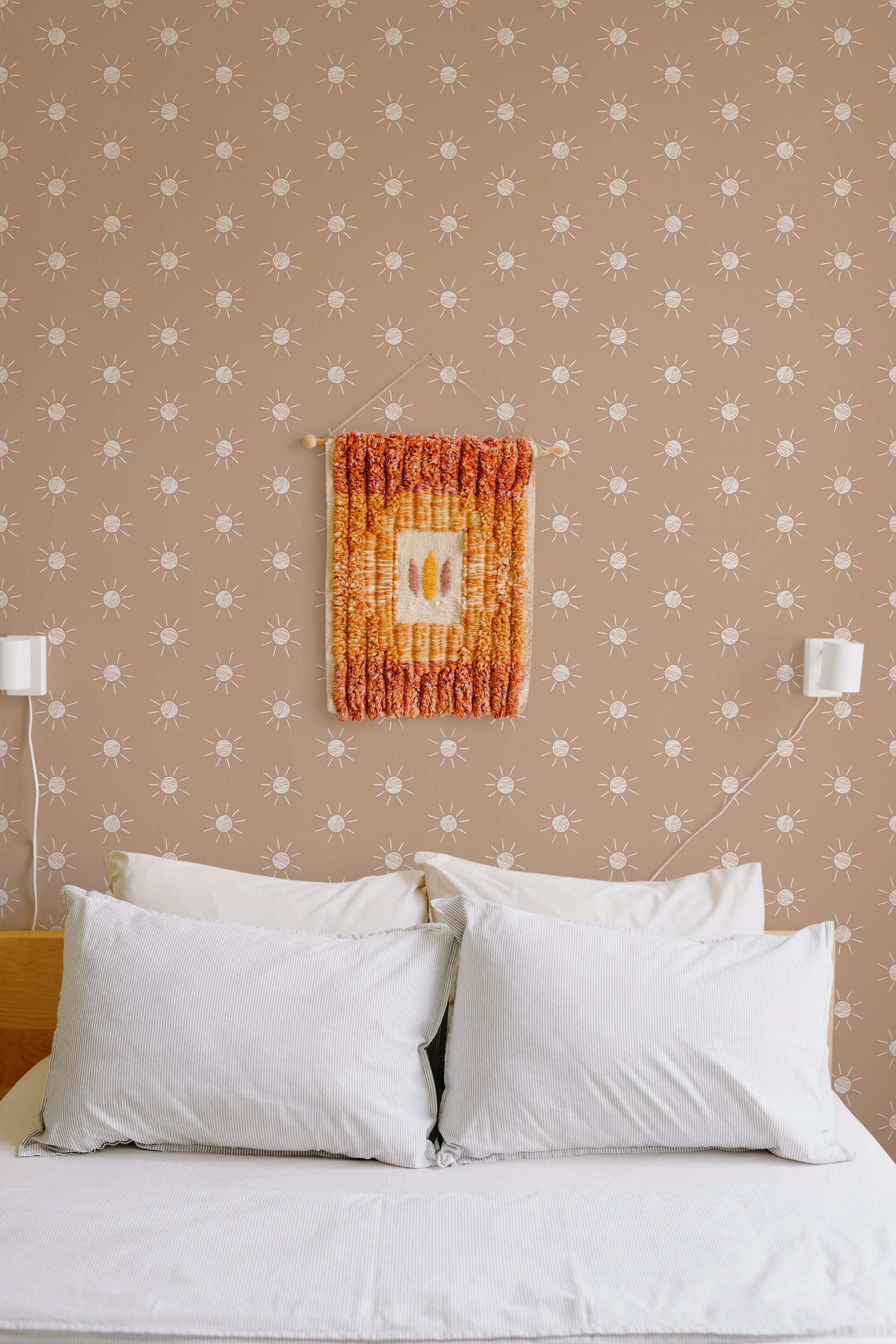 Self-adhesive wallpaper with a repetitive hand-drawn boho sun pattern in a neutral color scheme on a bedroom wall, above a bed with white pillows.