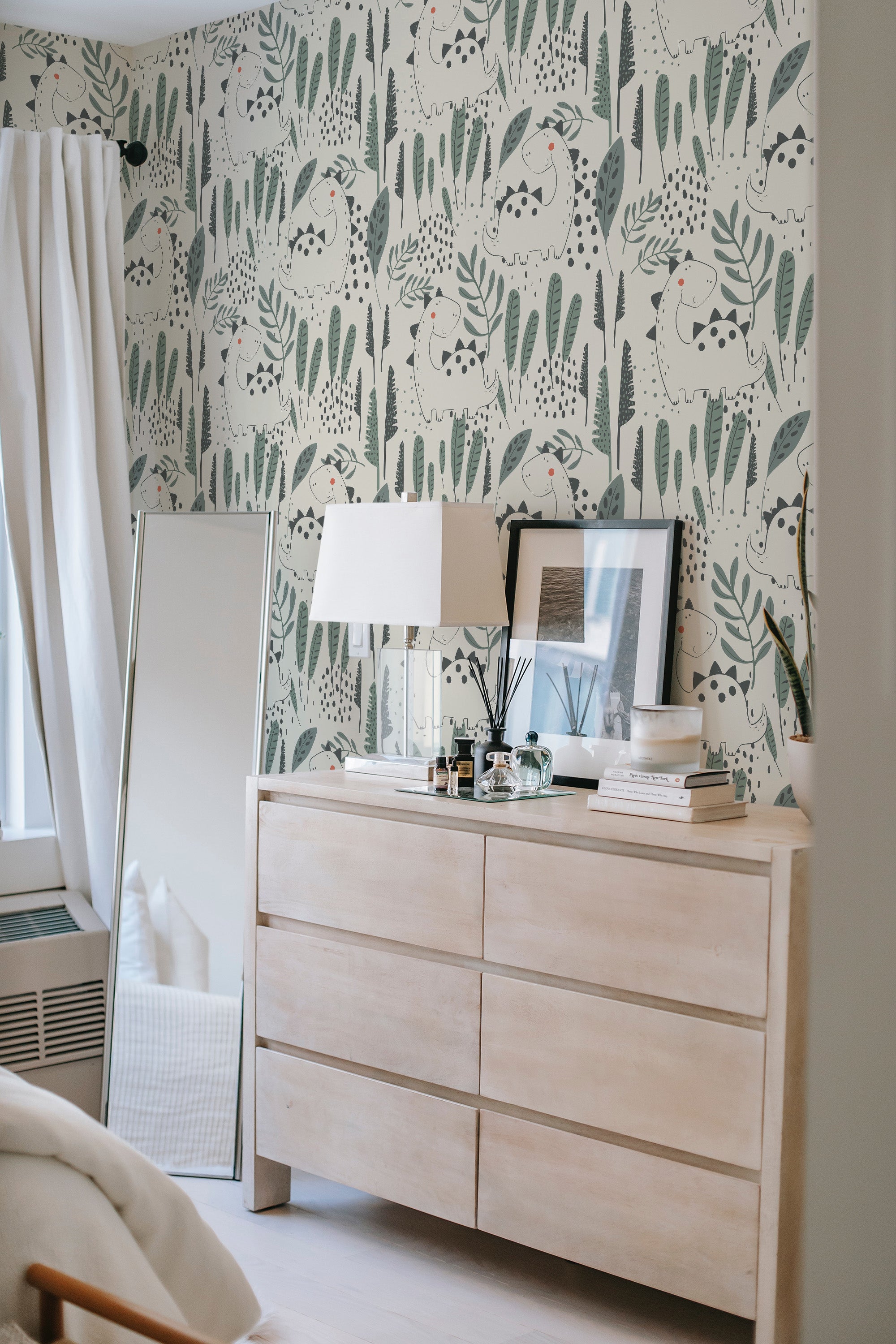A room with dinosaur-patterned wallpaper, including various dinosaurs in greens and blues amidst foliage on a light background, with a dresser and lamp in the foreground, giving a playful touch to traditional wallpaper.