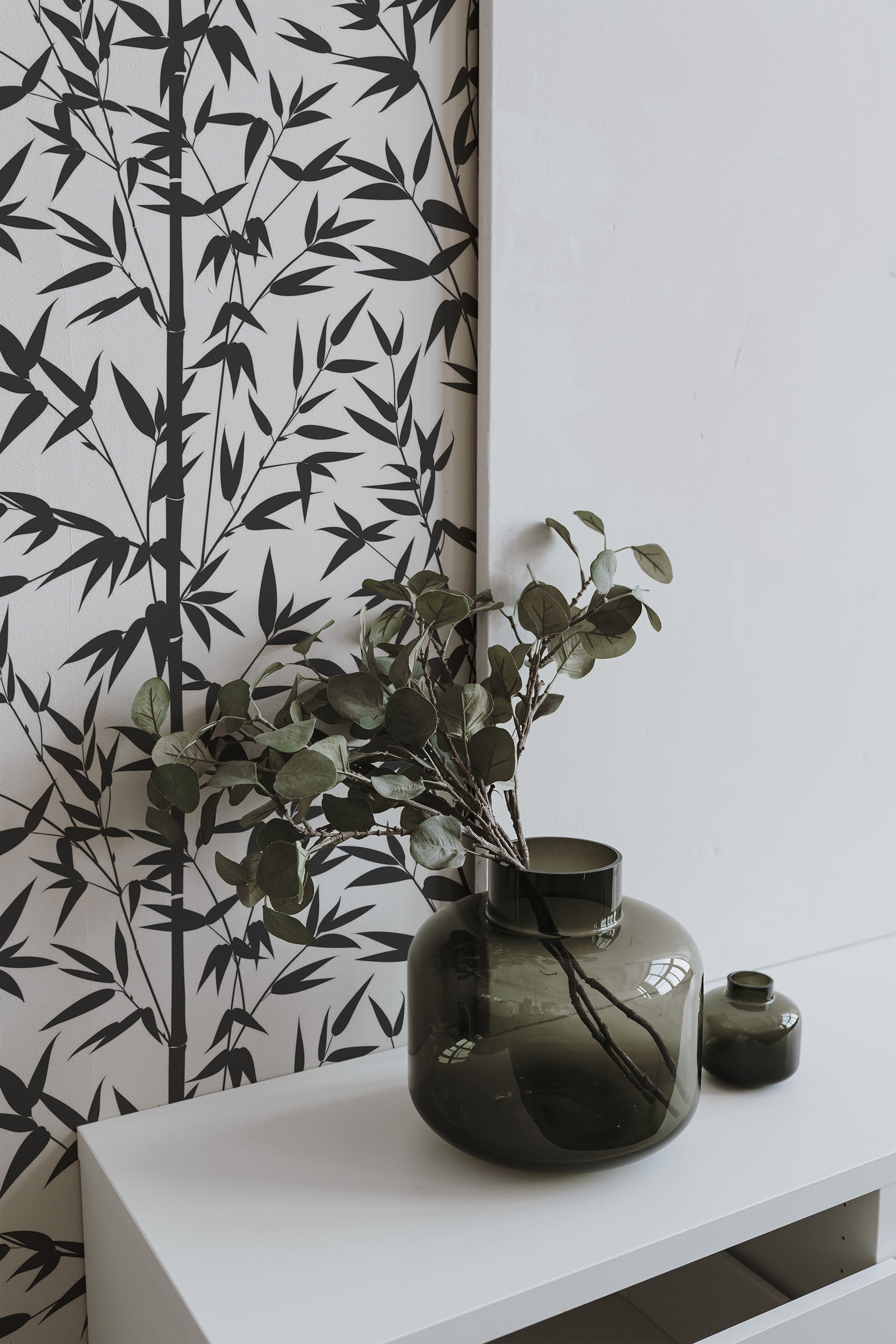 Self-adhesive wallpaper with black and white bamboo tree pattern on a room wall, complemented by a green vase with foliage on a white table.