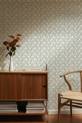 Self-adhesive wallpaper with a repeating simple leaf pattern in shades of green on a light background, applied on a wall above a wooden sideboard and beside a wooden chair.