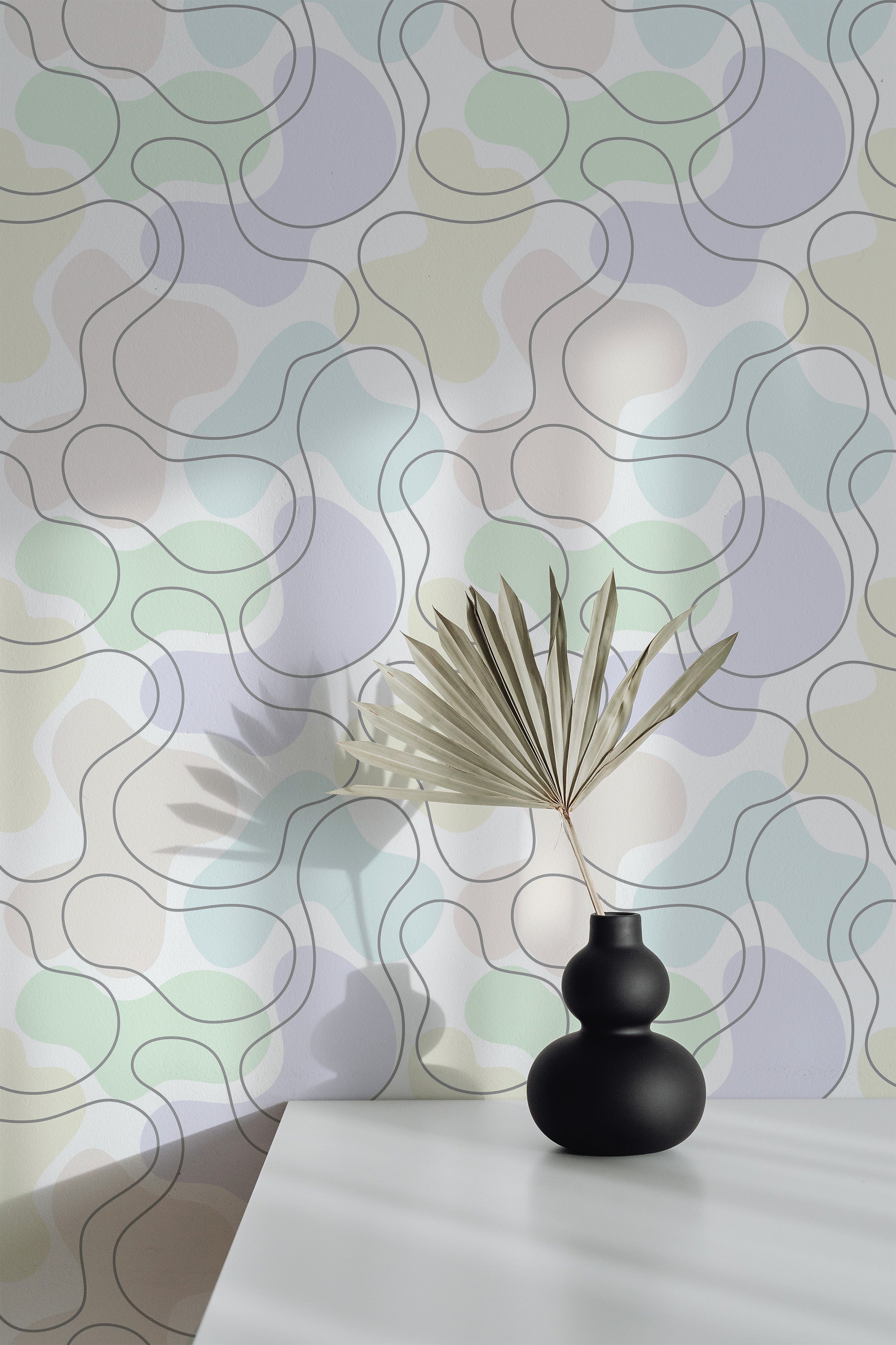 Self-adhesive wallpaper with an abstract pattern of interlocking shapes in pastel colors, complemented by a simple vase with a palm leaf on a white surface.