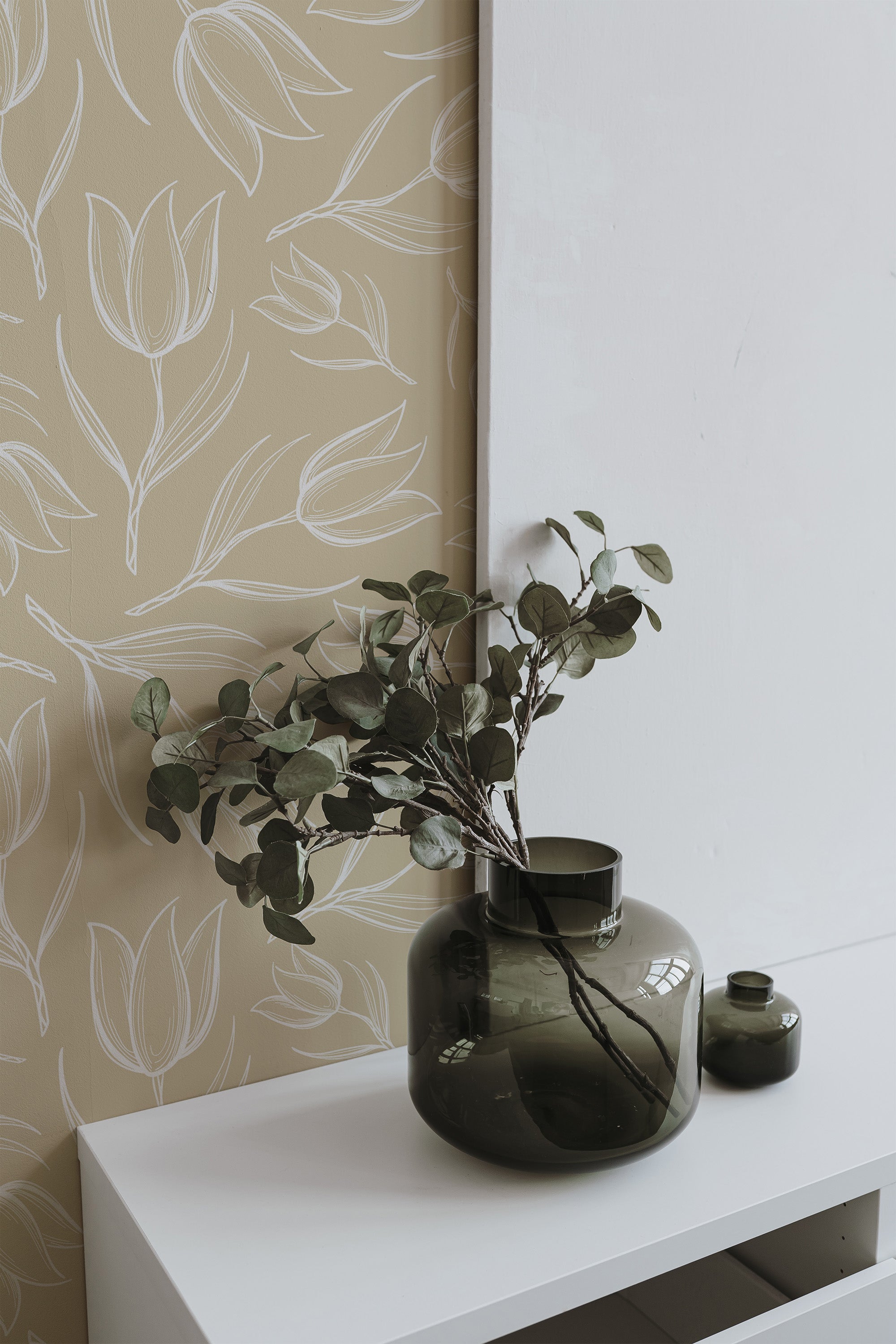 Self-adhesive wallpaper with a tulip pattern in shades of beige and cream on a wall, complemented by a green vase with foliage on a white shelf.