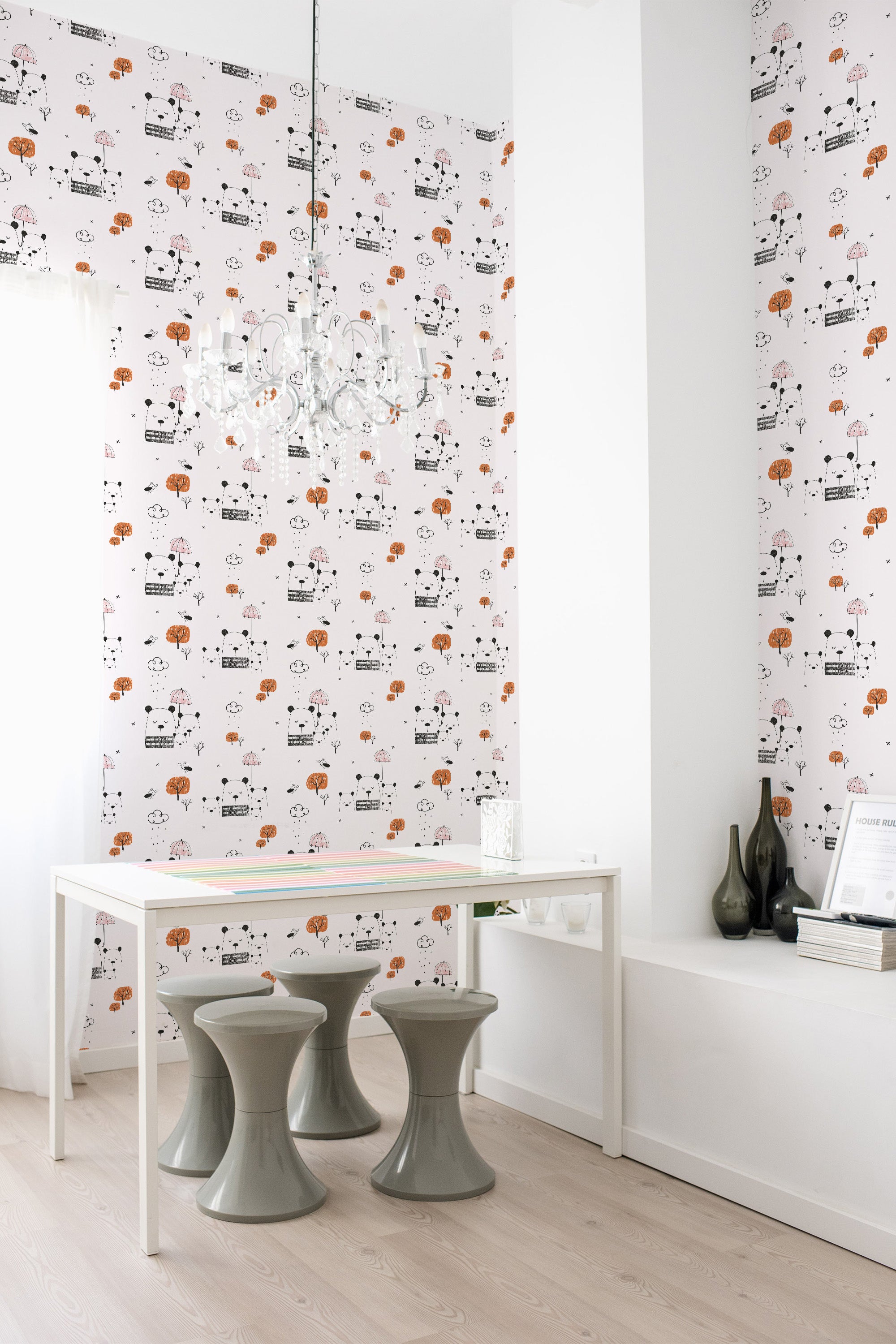 Wallpaper with playful bear illustrations in black, white, and brown, placed amid trees and small houses on a light background in a minimalist, modern room with a white table and grey stools. Traditional wallpaper.