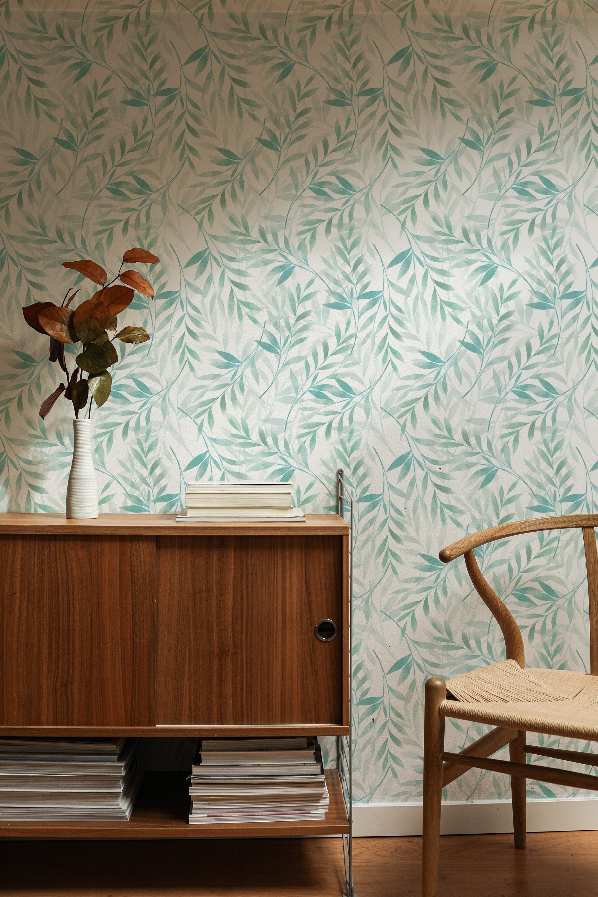 Self-adhesive wallpaper with a watercolor leaf design in soft shades of green and blue, creating a serene, nature-inspired look on the wall, paired with a wooden cabinet and a vase of dried flowers for a cozy, organic atmosphere in the room.