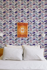 Self-adhesive herringbone wallpaper in shades of blue, gray, purple, and white with a modern, geometric pattern on the wall behind a neatly made bed.