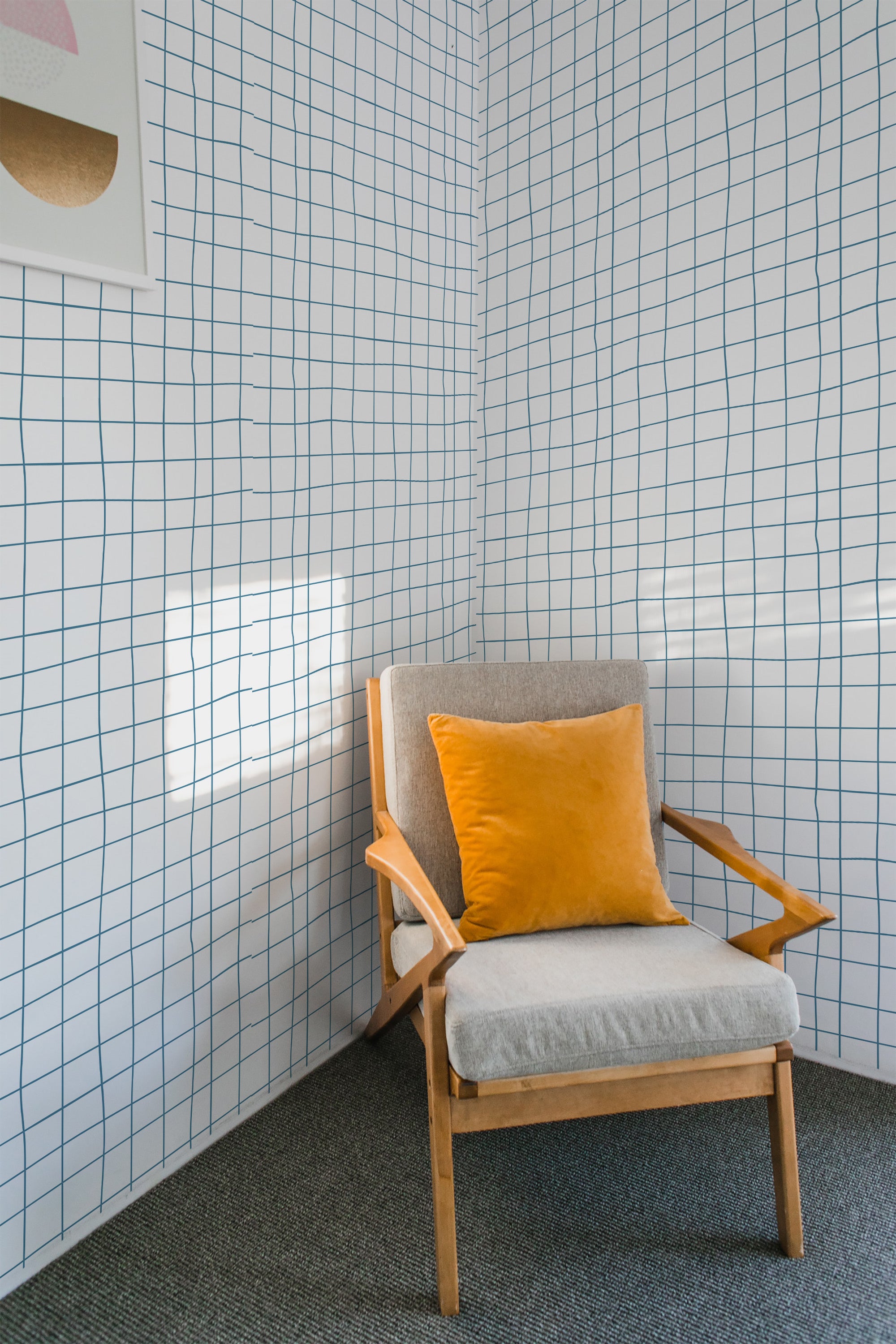 A room with Aesthetic grid wallpaper design, characterized by thin, uneven black lines forming irregular squares, over a white background. A wooden chair with a gray cushion and orange pillow sits in the corner. The wallpaper gives a contemporary twist to a traditional wallpaper.