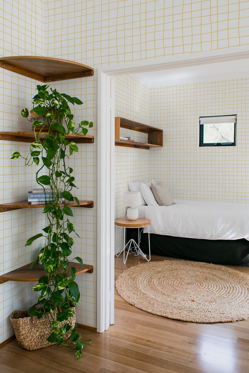 A room with walls covered in a white wallpaper adorned with a delicate yellow hand-drawn grid pattern, creating a minimalist yet engaging design. The grid lines are slightly uneven, adding a charming and organic touch to the overall aesthetic. The yellow color adds a subtle warmth against the white background, making the room feel cozy and inviting. The wallpaper complements the natural wood elements and houseplants in the room, evoking a relaxed atmosphere. Traditional wallpaper.