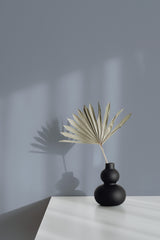 Self-adhesive solid color wallpaper in a soft, muted grey shade covering the wall behind a minimalist black vase holding a dried palm leaf.