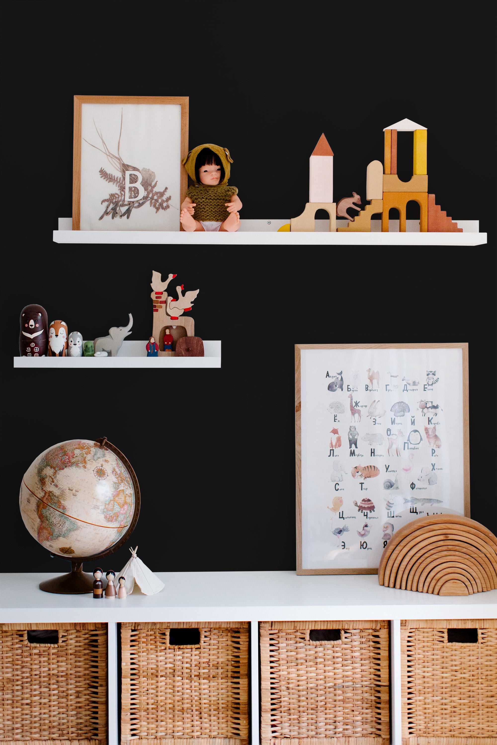 Self-adhesive bold solid black wallpaper adorning a wall behind white floating shelves and a white storage unit with wicker baskets. The wall is decorated with framed art, wooden toys, and a globe, creating a modern and playful atmosphere.